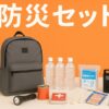 オレンジ色の背景に、防災セット（リュック、水、缶詰、懐中電灯、救急セット、手袋、ラジオ、乾電池）が整然と並べられている様子。