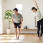 中高年の日本人男性と若い日本人女性が、明るくモダンな室内で、腰痛や姿勢改善のためにストレッチや軽い運動をしている様子を捉えた写真。