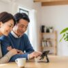 50代の日本人夫婦がリビングでスマートフォンスタンドを使い、笑顔でスマホを操作している様子。自然光の差し込む明るい部屋で快適なデジタル生活を楽しんでいるシーン